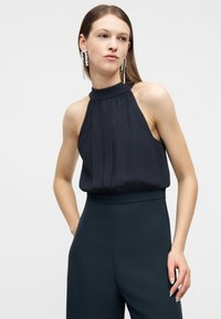 Γυναίκα που φοράει αμάνικο σκούρο τοπ με λαιμόκοψη halter και ταιριαστά ψηλόμεσα παντελόνια, με μακριά σκουλαρίκια με κρυστάλλινες σταγόνες, ποζάροντας μπροστά σε απλό φόντο.