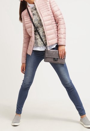 Hellrosa gepolsterte Jacke, grauer Pullover mit Blumenmuster, blaue Skinny-Jeans, silberne Sneakers und eine graue Umhängetasche. Lässiges Outfit und entspannte Pose.