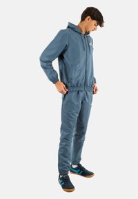 Chándal ligero azul con puños y cinturilla elásticos. Presenta una chaqueta con capucha y pantalones a juego. Se lleva con zapatos negros con acentos azules.