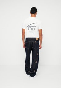 Hvit T-skjorte med blå grafikk på ryggen, kombinert med mørkeblå denimjeans med broderte krone-mønstre og hvite sneakers.