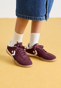 Burgundi velúr sportcipő fehér részletekkel és csillaglogóval, texturált gumitalppal és szövetfűzőkkel, fehér bordás zoknival viselve.