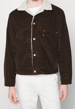 Homme portant une veste en velours côtelé brun foncé avec un col sherpa blanc et des boutons argentés, assortie à un pantalon blanc.