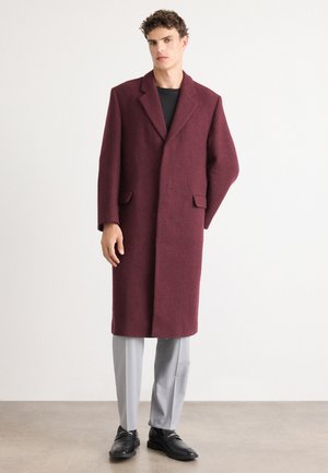 Abrigo de lana burdeos con cuello muesca, dos bolsillos frontales y un diseño oversized, combinado con pantalones gris claro y zapatos negros.