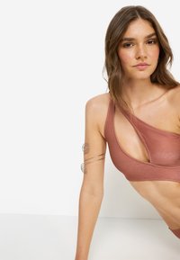 Αθλητικό σουτιέν σε χάλκινο χρώμα με έναν ώμο, σχέδιο με ανοίγματα, υφή υφάσματος και λεπτομέρεια με ασημένιο σπειροειδές κοσμηματικό βραχιόλι.