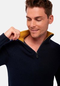 Sweat-shirt zippé bleu marine avec un col demi-hauteur ; col intérieur en jaune moutarde. Tissu texturé, avec une fermeture éclair.