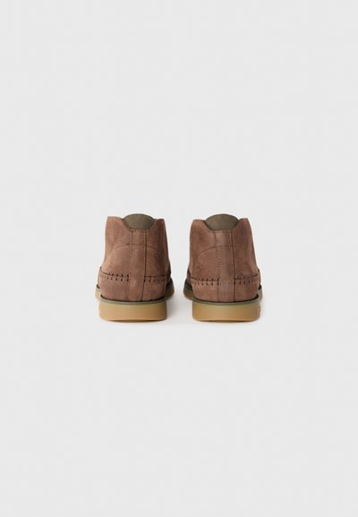 Scarpe in suede marrone con profilo basso, dettagli cuciti e suole in gomma. Presentano una parte posteriore arrotondata e una fodera interna in contrasto.