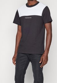 Camiseta de manga corta en blanco y negro con cuello redondo, con el logo "Calvin Klein Jeans" en el pecho. Tela de algodón, corte relajado.