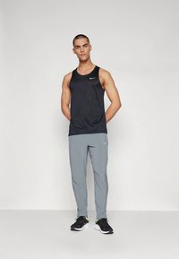 Hombre de pie con las manos detrás de la espalda, vistiendo una camiseta sin mangas negra de Nike, pantalones grises de Nike y zapatillas negras de correr, sobre un fondo liso.