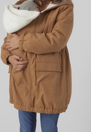 Personne portant une veste marron tenant un bébé enveloppé dans un bonnet en maille gris et une couverture blanche près de sa poitrine.