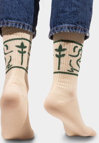 Beżowe skarpetki w prążki z zielonymi graficznymi wzorami, w tym roślinami i postaciami, widocznymi w górnej części. Noszone z dżinsami z podwijanymi nogawkami.
