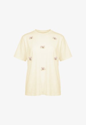Kremowy T-shirt z krótkim rękawem z sześcioma małymi różowymi haftowanymi kokardkami rozmieszczonymi z przodu. Brak widocznych logo ani innych wzorów.
