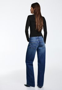 Donkerblauwe denim jeans met hoge taille, een relaxte pasvorm en achterzakken met zilverkleurige studs. Zwart shirt met lange mouwen gedragen.