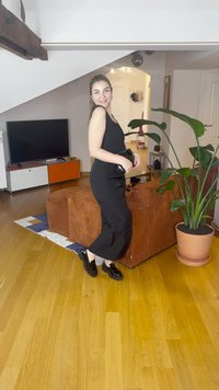 Svart kjole med en tettsittende silhuett og utvidet nederdel; brukt med svarte loafers. Bakgrunnen inkluderer en brun ribbet sofa og en potteplante.