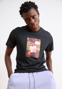 Schwarzes Baumwoll-T-Shirt mit einem zentralen Foto eines Mannes, der einen Basketball hält. Er trägt ein rot-weißes Trikot mit dem Schriftzug "BULLS". Glatte Textur.