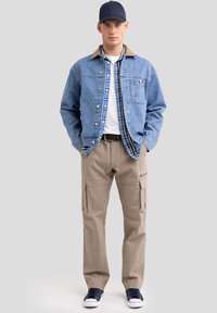 Denim jakna z rjavim ovratnikom, preko modre karirane srajce, bež cargo hlače in mornarsko modri športni čevlji. Odlika sproščenega slojevitkega videza.