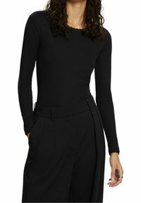 Personne portant un haut noir cintré à manches longues avec bordure en dentelle et un pantalon noir taille haute à jambes larges, les mains partiellement visibles.