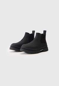 Botas Chelsea pretas feitas de material sintético liso, com um amplo painel lateral elástico, uma alça de puxar e uma sola de borracha espessa com sulcos.