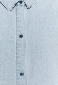 Chemise en jean bleu clair avec un col, six boutons sur le devant et une texture douce avec des variations subtiles de teinte.