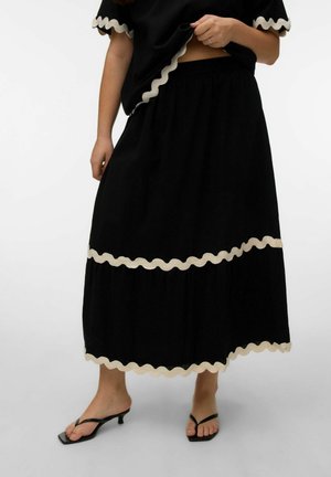 Kvinde iført sort midi-nederdel og top med beige bølget kant, kombineret med sorte sandaler med kitten-hel, foran en hvid baggrund.