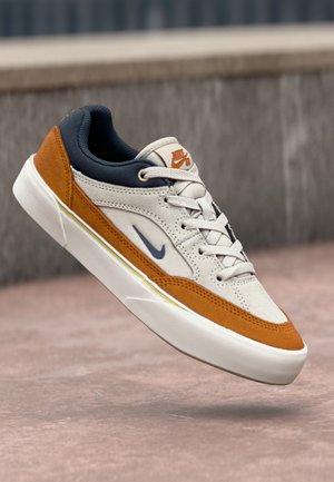 Nike SB sneaker featuring a cream fabric upper, orange suede accents, a navy blue collar, and a white rubber sole with a sleek design.
