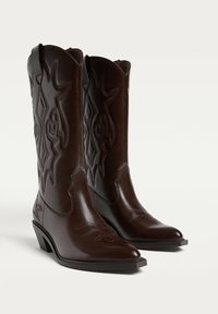 Braune Leder-Cowboystiefel mit spitzen Zehen, dekorativen Nähten, verjüngten Absätzen und gewellten oberen Kanten, die aufwendige geprägte Muster aufweisen.