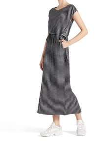 Kvinna i en svart och vit horisontellt randig midiklänning med sidofickor, ihop med vita sneakers, som står mot en vit bakgrund.