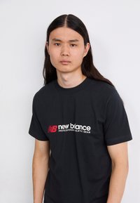 Sort bomulds T-shirt med rund halsudskæring, prydet med et hvidt og rødt "new balance" logo og teksten "PROFESSIONAL ATHLETIC GEAR" på fronten.