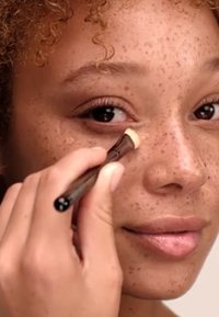 Pennello per correttore tenuto da una mano che applica crema sotto l'occhio, con un manico in legno scuro e un prodotto beige chiaro che si fonde con una pelle cosparsa di lentiggini.