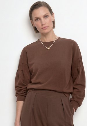 Donna con capelli corti che indossa una maglia marrone a maniche lunghe e pantaloni con maniche a pieghe e una collana di perle con un ciondolo d'oro.