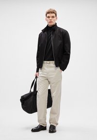 Μαύρο μπουφάν bomber πάνω από έναν μαύρο ζιβάγκο, μπεζ παντελόνι, μαύρα δερμάτινα παπούτσια, κρατώντας μια μεγάλη μαύρη τσάντα ταξιδιών. Μινιμαλιστικός σχεδιασμός.