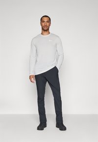 Helles graues Langarmshirt mit kleinem Logo, kombiniert mit dunklen Hosen und schwarzen Schuhen, vor einem einfarbigen Hintergrund stehend.