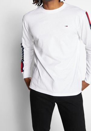 Mężczyzna ubrany w białą koszulę z długim rękawem, z czerwono-biało-niebieskimi paskami na rękawach i małym logo na piersi, w połączeniu z czarnymi spodniami.