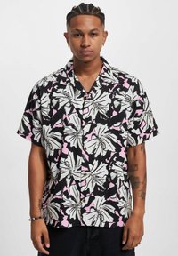 Camisa de manga corta con fondo negro, patrón floral en blanco y rosa, y botones marrones. Hecha de una tela ligera y texturizada.