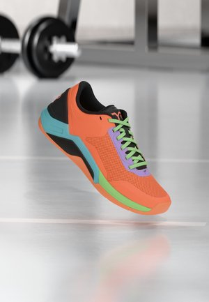 Jasně oranžová sportovní bota se zelenými tkaničkami, fialovým jazykem a modrými a černými akcenty, zavěšená nad lesklou podlahou posilovny s činkami v pozadí.