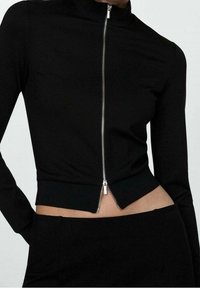 Sort cropped jakke med høj krave, lange ærmer, lynlås foran og tætsiddende design. Materialet ser glat ud med en let glans.
