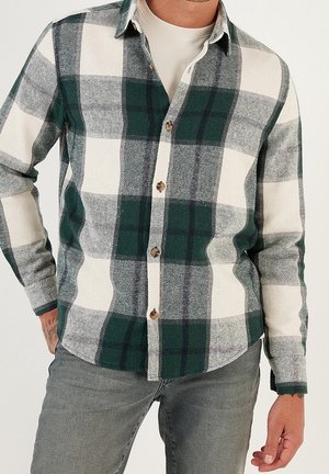 Homme portant une chemise à carreaux verte, blanche et noire par-dessus un t-shirt beige et un jean gris, debout avec une main derrière le dos.