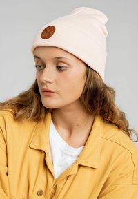 Jasnoróżowa dziana czapka z mankietem i okrągłą, brązową skórzaną metką z wytłoczonym wzorem. Miękka tekstura i gładkie wykończenie.