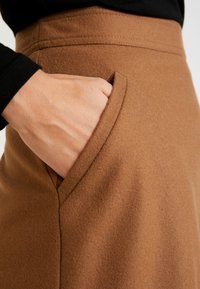 Main dans la poche latérale d'une jupe en laine marron portée avec un haut noir à manches longues.