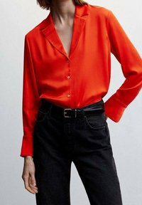 Camicetta rossa in satin con bottoni, colletto a risvolto e maniche lunghe, indossata con jeans neri a vita alta e una cintura nera. Texture liscia.