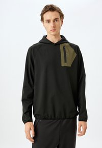 Sudadera negra con capucha que presenta un bolsillo de parche en color oliva contrastante con cremallera. El material es suave y el diseño incluye una cintura ajustable.