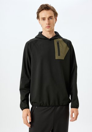 Sudadera negra con capucha que presenta un bolsillo de parche en color oliva contrastante con cremallera. El material es suave y el diseño incluye una cintura ajustable.