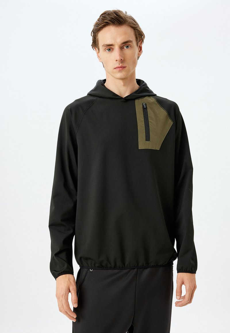 Sudadera negra con capucha que presenta un bolsillo de parche en color oliva contrastante con cremallera. El material es suave y el diseño incluye una cintura ajustable.