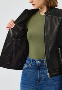 Chaqueta de cuero negra con cierre de cremallera y un cuello texturizado, llevada sobre una blusa verde; cuenta con un forro interior estampado y jeans azules.