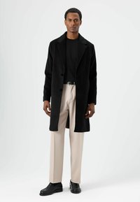 Zwarte wollen jas met een ingeslagen kraag en knoopsluiting; gedragen over een zwart overhemd en lichtbeige broek, gecombineerd met zwarte schoenen.