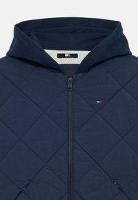 Tamsiai mėlynas quilted tipo švarkas su kapišonu, priekiniais užtrauktukais ir mažo logotipo etikete su raudonais, baltos ir mėlynos spalvos akcentais šalia kaklo.