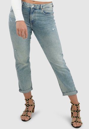 Lichtblauwe spijkerbroek met versleten details, rechte snit, opgerolde manchetten, gecombineerd met zwarte sandaaltjes met bandjes en gouden accenten.
