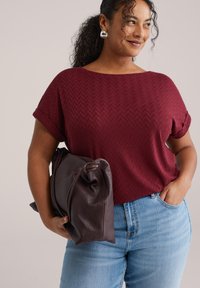 Weinrotes Kurzarmshirt mit Chevron-Muster, kombiniert mit hellblauen Jeans und einer braunen, lässigen Ledertasche. Silberne Creolen runden den Look ab.