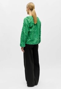 Femme avec une queue de cheval blonde portant un chemisier vert à motifs floraux texturés et un pantalon noir ample, debout de dos sur un fond clair uni.