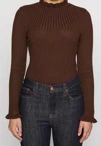 Haut marron à manches longues côtelées avec un col montant à volants, associé à un jean foncé avec des poches traditionnelles et une fermeture à boutons.