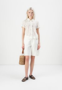 Camisa blanca de encaje de manga corta con cuello, combinada con pantalones cortos de mezclilla blanca. Los accesorios incluyen un bolso de palma tejido y zapatos planos de estampado de leopardo.